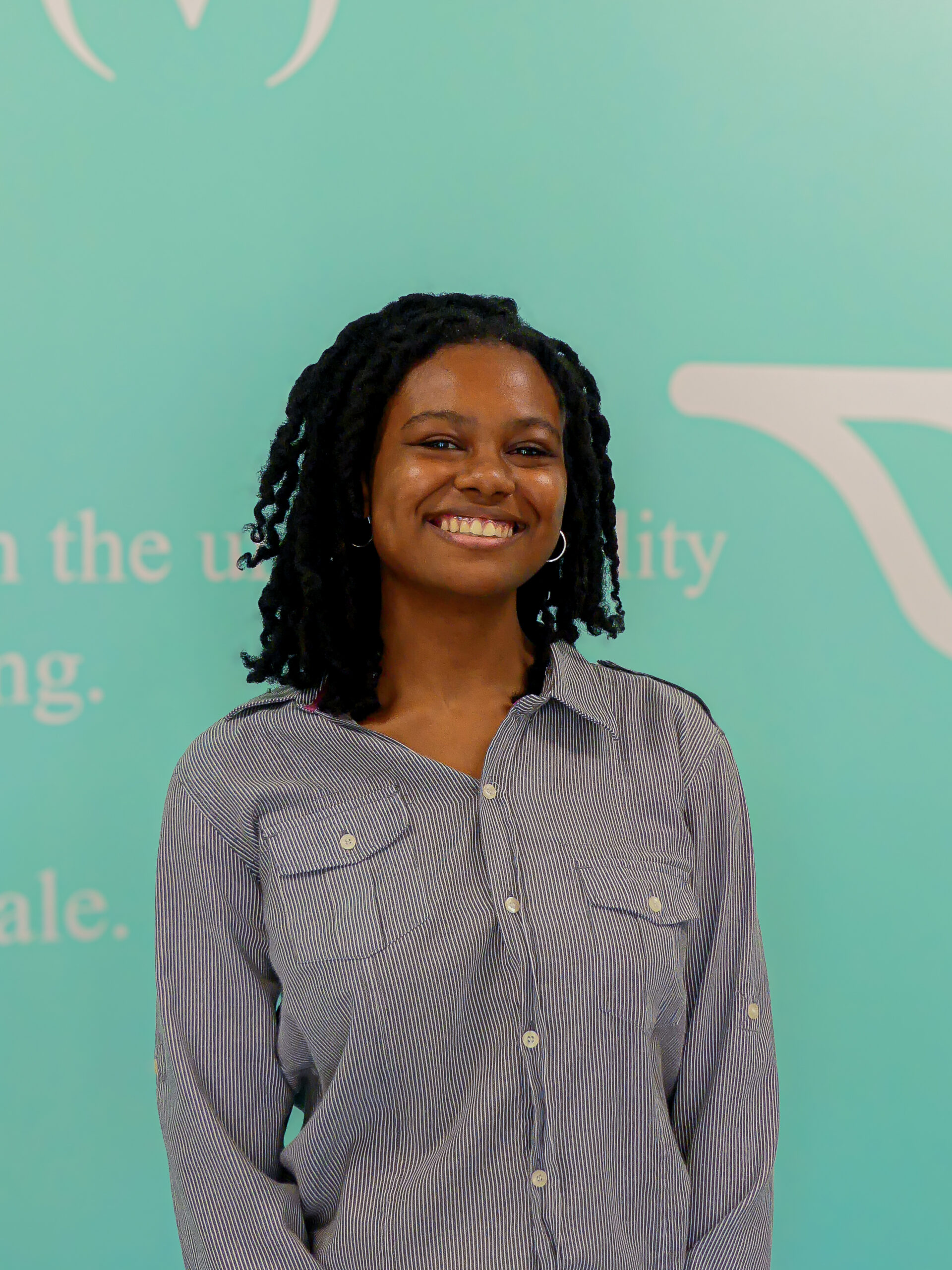 Smiling woman with braided hair wearing a striped button-down shirt, standing in front of a teal backdrop.