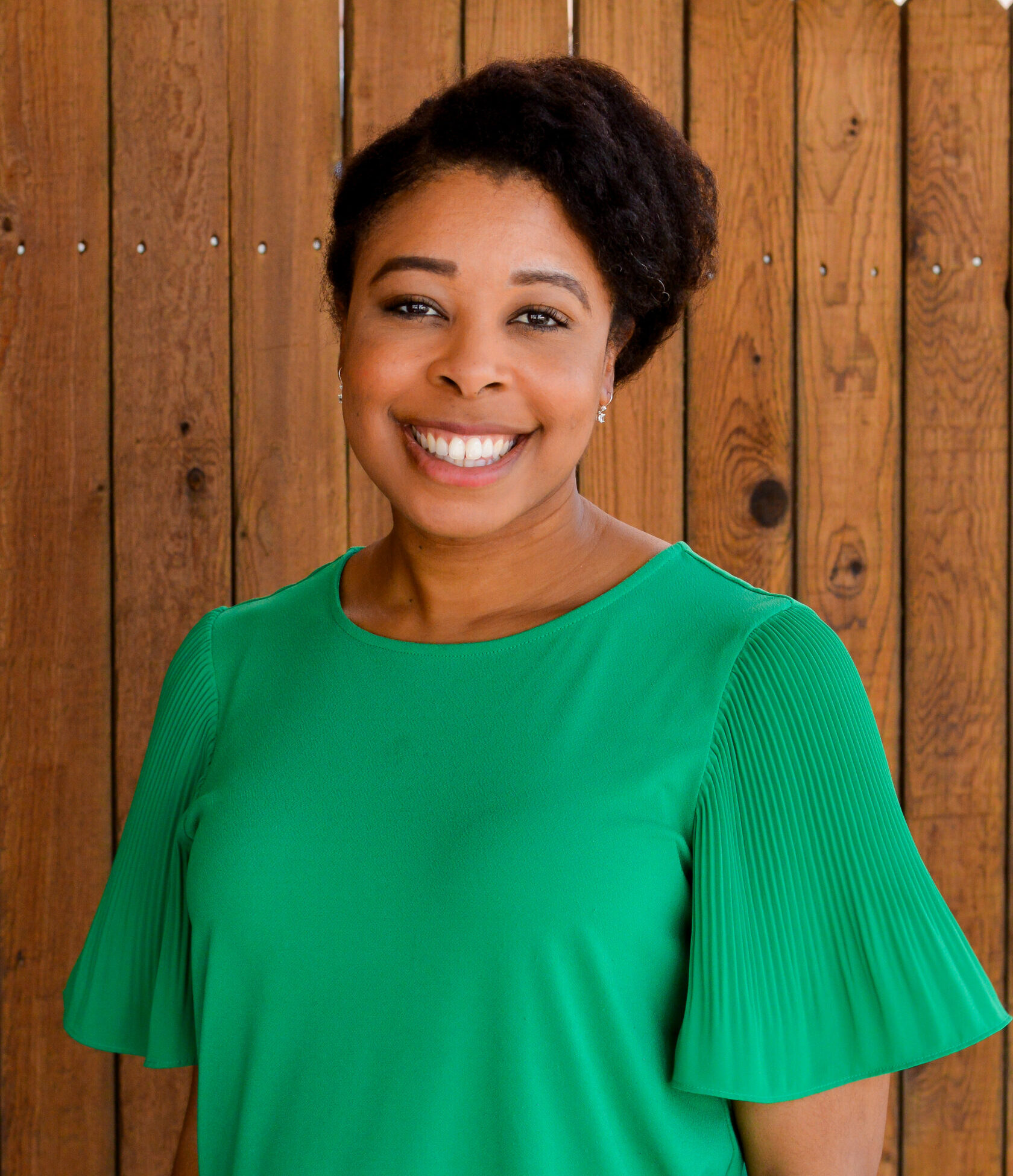 Smiling woman in a bright green dress, standing in front of a wooden fence.
